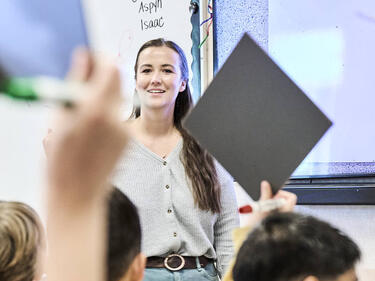 Teacher standing at the front of a classroom while students seated at desks raise whiteboards and hands to participate in a lesson. A whiteboard and learning materials are visible in the classroom background.
