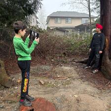 One student taking a photo of another student who is posing and leaning against a tree